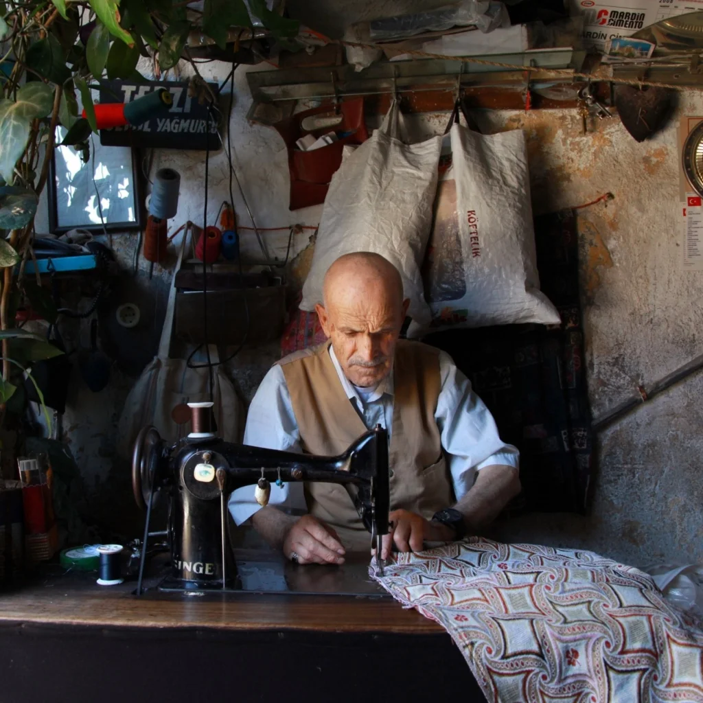 Elderly tailor sewing fabric with vintage machine in rustic workshop.