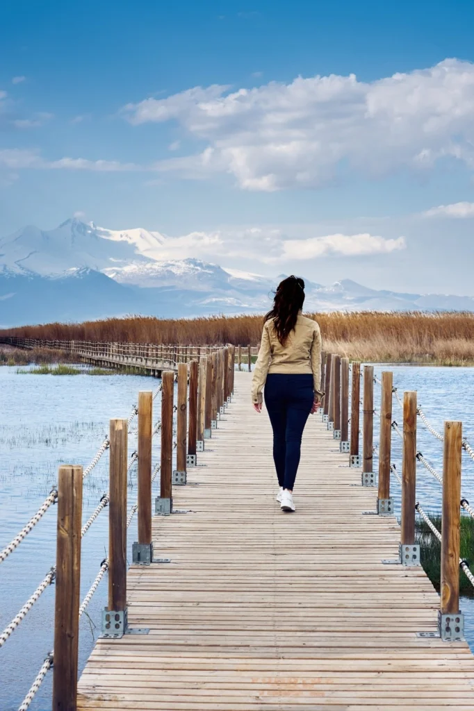 Woman walking on a wooden pier towards mountains under a blue sky.