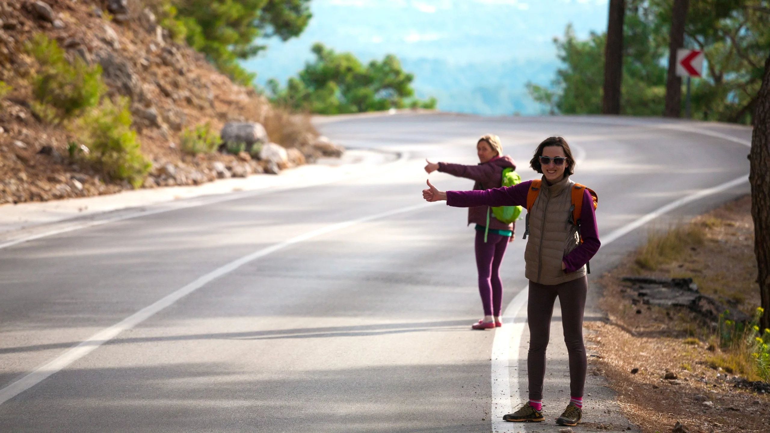 Two women hitchhiking on a scenic road surrounded by trees.