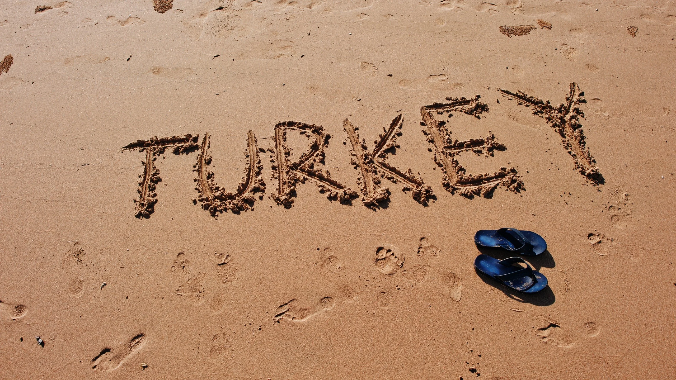 The word 'Turkey' written in sand with blue sandals nearby.